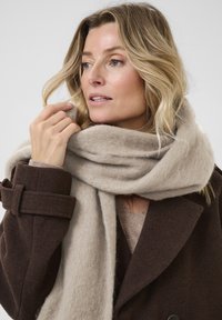 Beige Wollschal, der um den Hals drapiert ist, weiche Textur, kombiniert mit einem dunkelbraunen Mantel mit Gürtel und großen Knöpfen.