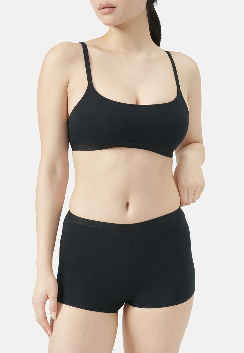 Γυναίκα που φοράει μαύρο αόρατο σουτιέν και αντίστοιχα μποξεράκια, σε απλό ανοιχτό φόντο.