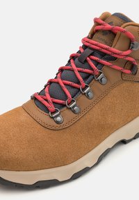 Brune semskede tursko med røde snøring, en teksturert svart tunge, metalliske snørehull og en kontrastrik beige såle med grep mønster.
