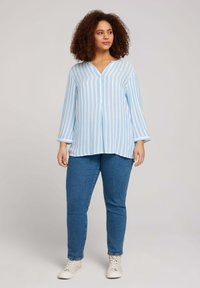 Camisa de manga larga a rayas en azul claro y blanco con escote en V, detalles de botones y un corte relajado, combinada con jeans de color azul oscuro y zapatillas blancas.