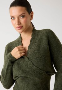 Pulover tricotat de culoare verde măslin, cu decolteu adânc în V, textură cu nervuri, mâneci lungi și un guler cu margini zimțate, având o siluetă ajustată.