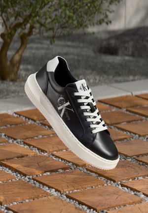 Zwart-witte Calvin Klein sneaker met witte veters, logo op de zijkant en tong, rustend op een houten tegel en grind buiten.
