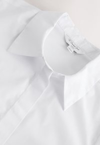Col de chemise blanche nette avec boutons cachés et une étiquette affichant "The Comfort Shirt" dans un style flat lay.