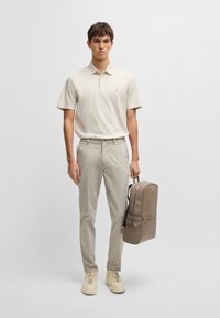 Beige polo skjorte, lyse beige bukser og beige sneakers. Holder en brun ryggsekk med en glatt overflate og justerbare stropper.