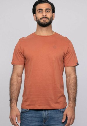 Homme barbu portant un t-shirt à manches courtes de couleur rouille et un jean bleu, debout devant un fond clair uni.