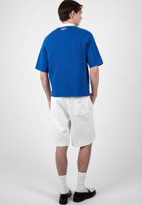 Hombre con una camisa azul de manga corta con estampado, pantalones cortos blancos con bolsillo trasero, calcetines blancos y zapatos de vestir negros, de pie de espaldas.