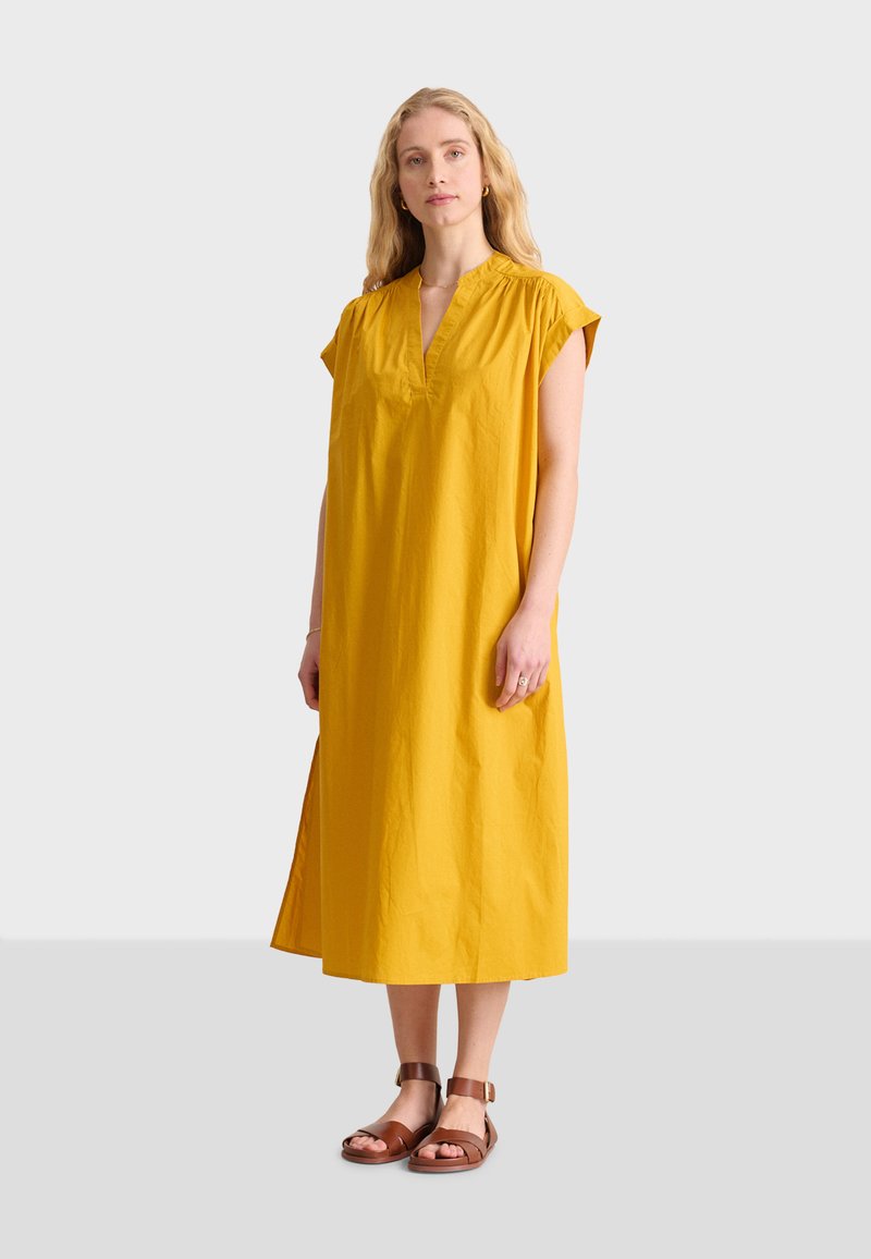 Gelbes Baumwollkleid mit kurzen Cap-Ärmeln, V-Ausschnitt, leichtem Stoff und seitlichen Schlitzen; kombiniert mit braunen Riemchensandalen.