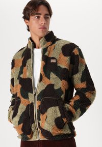 Giacca in pile con un motivo in camouflage marrone, verde e beige, chiusura a zip, colletto alto e tasche laterali con una superficie texturizzata.