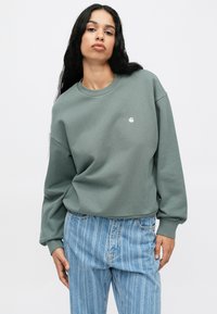 Zöld pulóver, kerek nyakkal és bordázott mandzsettával, a mellkason egy kis logóval. Magas derekú, világoskék csíkos farmernadrággal párosítva.