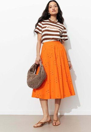 Femme portant un t-shirt rayé marron et blanc rentré dans une jupe midi orange vif à œillets, tenant un sac souple marron texturé, et portant des sandales beige.