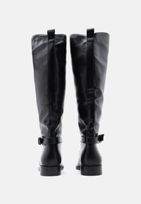Μαύρες δερμάτινες μπότες μέχρι το γόνατο με λεία υφή, στρογγυλεμένη μύτη, πλευρικό λουράκι με αγκράφα και χαμηλό στιβαρό τακούνι. Μινιμαλιστικός σχεδιασμός.