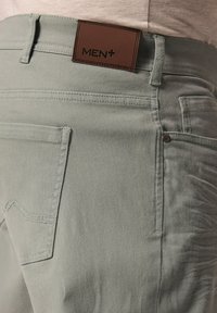 Vihreät farkut, joissa on ruskea nahkainen etiketti, jossa lukee "MEN+", perus-taskut ja kontrastiväriset ompelut. 