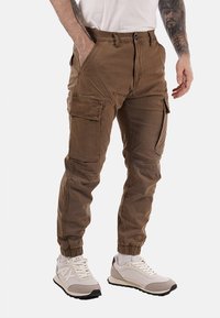 Pantaloni cargo in un tessuto marrone neutro, con numerose tasche, polsini elasticizzati e una vestibilità rilassata. Abbinati a sneakers dai toni neutri.