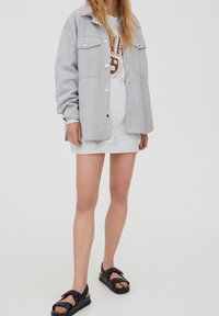Lys grå oversized jakke med knaplukning, to brystlommer, lagdelt over en grå sweatshirtkjole. Sorte sandaler med stropdetaljer.