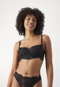 Melnā mežģīņu bralette ar caurspīdīgu materiālu, ziedu rakstiem, apakšveļas atbalstu un regulējamām siksnām. Saskaņota ar atbilstošām mežģīņu bikini apakšbiksēm.