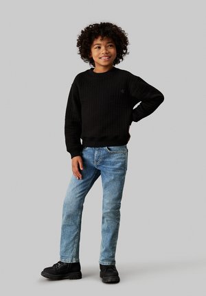 Maglione nero a coste, jeans azzurri chiari e stivali neri stringati. Il maglione presenta un piccolo logo sul petto. La texture sembra morbida.