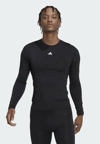 Svart langermet treningstopp laget av glatt, elastisk stoff. Har en liten hvit Adidas-logo på brystet. Passformen er tett inntil kroppen.