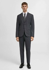 Sivi odijelo s oštrim reverima, zatvaranje na jedan gumb, usklađene hlače i bijela košulja, u paru s tamnom kravatom i crnim cipelama.