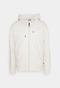 Haut de survêtement zippé crème clair avec un motif texturé. Il comprend une capuche à cordon et des poignets côtelés, fabriqué à partir d'un tissu doux.