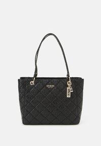 Sac cabas matelassé noir en cuir synthétique, avec une plaque logo dorée, deux poignées robustes et une étiquette porte-bonheur, au toucher lisse.