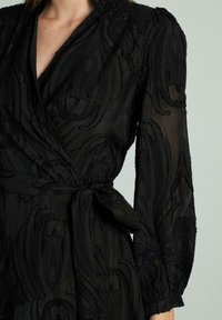 Schwarzes Wickelkleid aus strukturiertem Stoff mit einem aufwendigen Paisleymuster, langen Ärmeln und einem gebundenen Taillendetail.