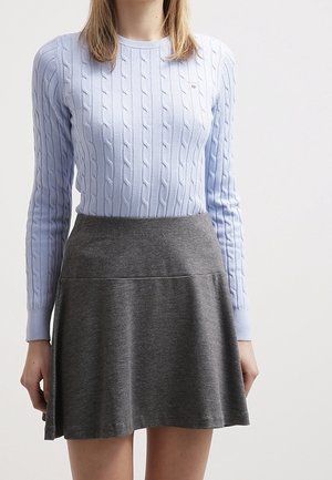 Kobieta ubrana w jasnoniebieski sweter warkoczowy wpuszczony do wysokiej taliowanej, szarej spódnicy typu A, stojąca na jednolitym tle.