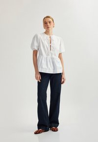 Blouse blanche à manches bouffantes avec un décolleté en clé et un détail à nouer, associée à un jean bleu marine évasé et des ballerines rouges ornées.