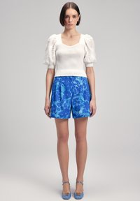 Top bianco in maglia con maniche a palloncino, abbinato a pantaloni corti blu con motivo floreale e scarpe blu. Texture lisce e colori a contrasto.