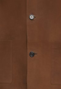 Brown suede jacket with two front pockets, featuring a round black button on the right side and clean stitching, smooth texture.