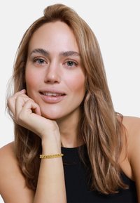 Bracciale in oro alla caviglia, con maglie intrecciate, design circolare e un finish lucido. Primo piano di una donna con capelli lunghi.