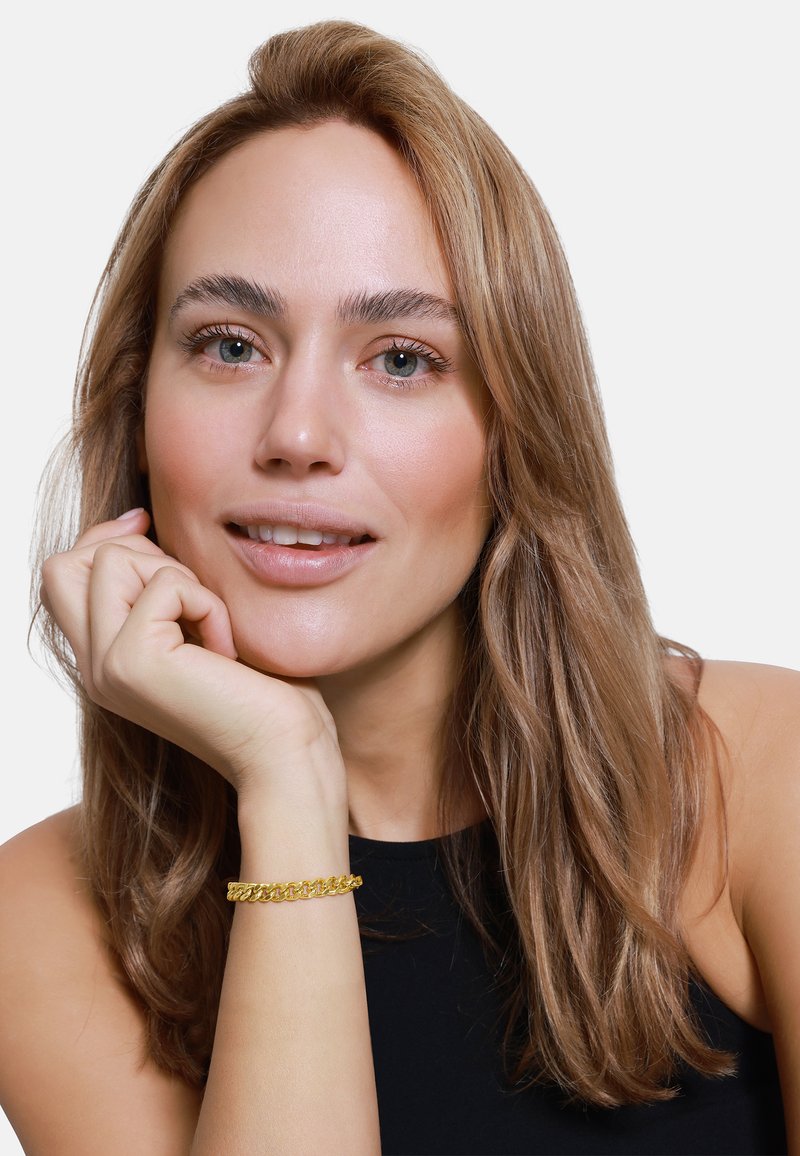 Bracciale in oro alla caviglia, con maglie intrecciate, design circolare e un finish lucido. Primo piano di una donna con capelli lunghi.