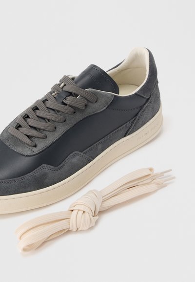 Γκρι δερμάτινο και καστόρινο αθλητικό παπούτσι με μπεζ ελαστική σόλα, διαθέτει γκρι κορδόνια και λείο υφή. Περιλαμβάνει ένα μπεζ κορδόνι δεμένο σε κόμπο στο πλάι.