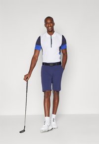 Pánský golfový outfit skládající se z bílého a námořnického polo trika s modrými detaily, doplněný námořnickými šortkami, černým opaskem a bílými golfovými botami se hroty.