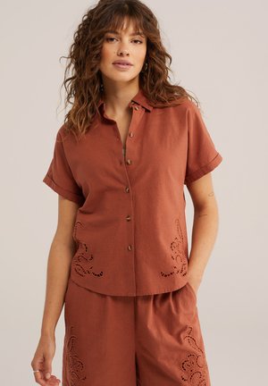 Frau trägt eine kurzärmelige, rostfarbene Knopfleiste-Bluse und passende Shorts mit dekorativen Ausschnittmustern, steht mit einer Hand in der Tasche.