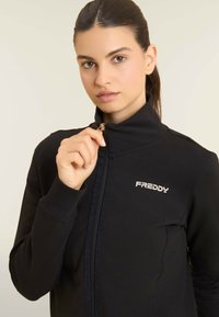 Giacca nera con zip realizzata in tessuto elastico, con colletto alto, zip frontale e logo bianco "FREDDY" sul petto.