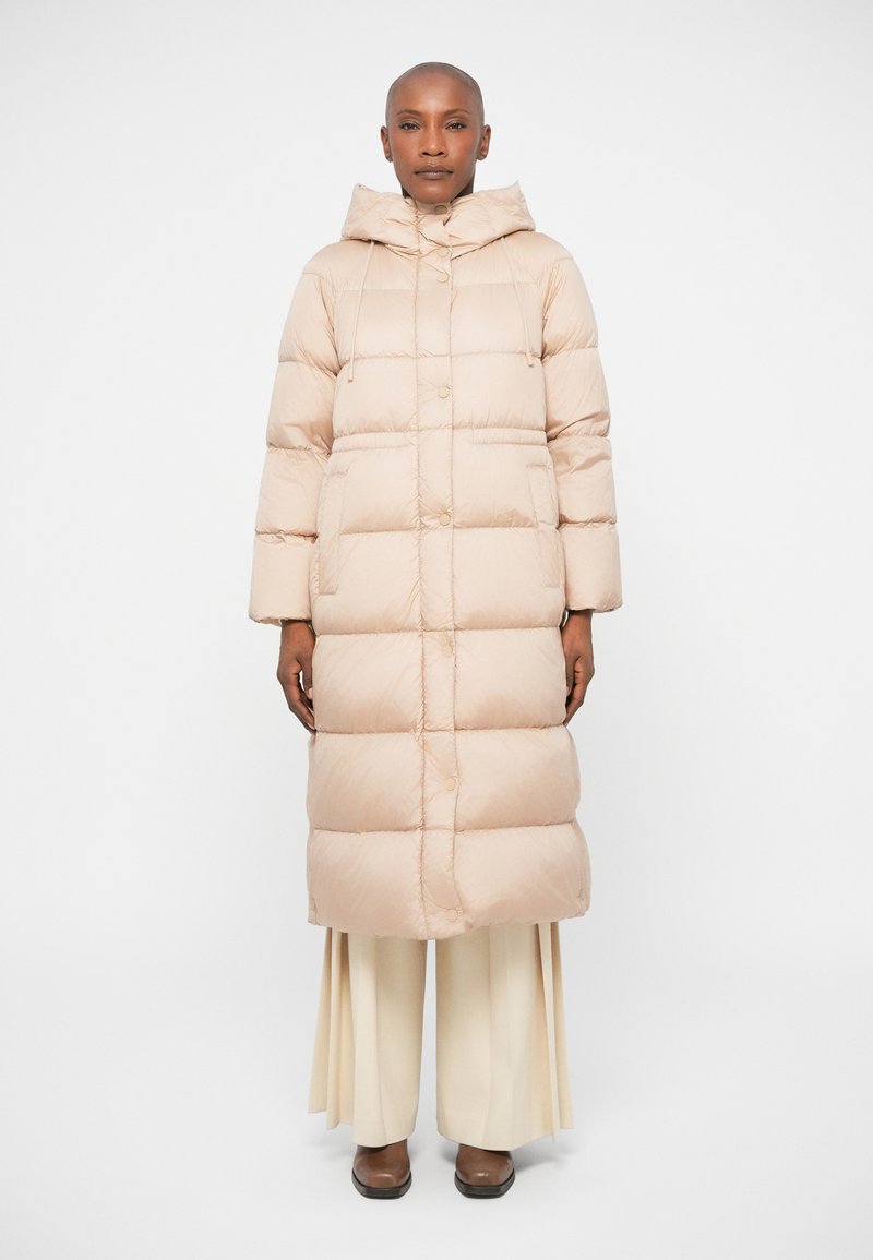 Beige puffertáska kapucnival, nagy vízszintes steppelésekkel, gombos elülső résszel és rugalmas állítható zsinórokkal. Tejfehér, bő szárú nadrággal viselve.