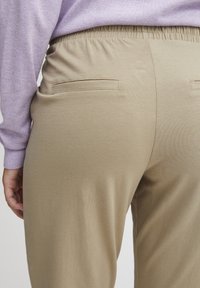 Beige bukser med elastisk liv med to baklommer. Stoffet ser mykt og glatt ut, vist sammen med en lys lilla topp.