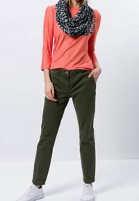 Maglietta a maniche lunghe color corallo, pantaloni verdi, sciarpa nera a fantasia, scarpe bianche. I pantaloni presentano tasche frontali e una vestibilità comoda. Tessuto morbido.