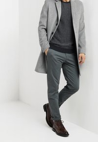 Šedý vlněný kabát přes černý texturační svetr, v kombinaci s tmavě zelenými kalhotami a hnědými koženými botami. Jednoduchý, dobře střižený design.