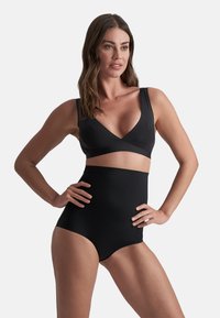 Donna che indossa un perizoma nero a vita alta e un reggiseno nero abbinato con spalline larghe, in piedi con le mani sui fianchi contro uno sfondo semplice.