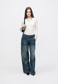 Mulher vestindo uma blusa bege de mangas compridas, jeans azuis de perna larga, sapatos pretos com bico fino, colar de corrente prateada, segurando uma mala preta de ombro.