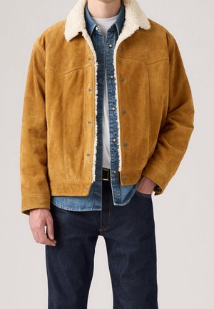 Mies pukeutunut sinapinväriseen nupukkinahkatakkiin, jossa valkoinen karvahuppuinen kaulus, sinisen farkkupaidan ja valkoisen aluspaitaan kanssa, yhdistetty tummansinisien farkkujen kanssa.