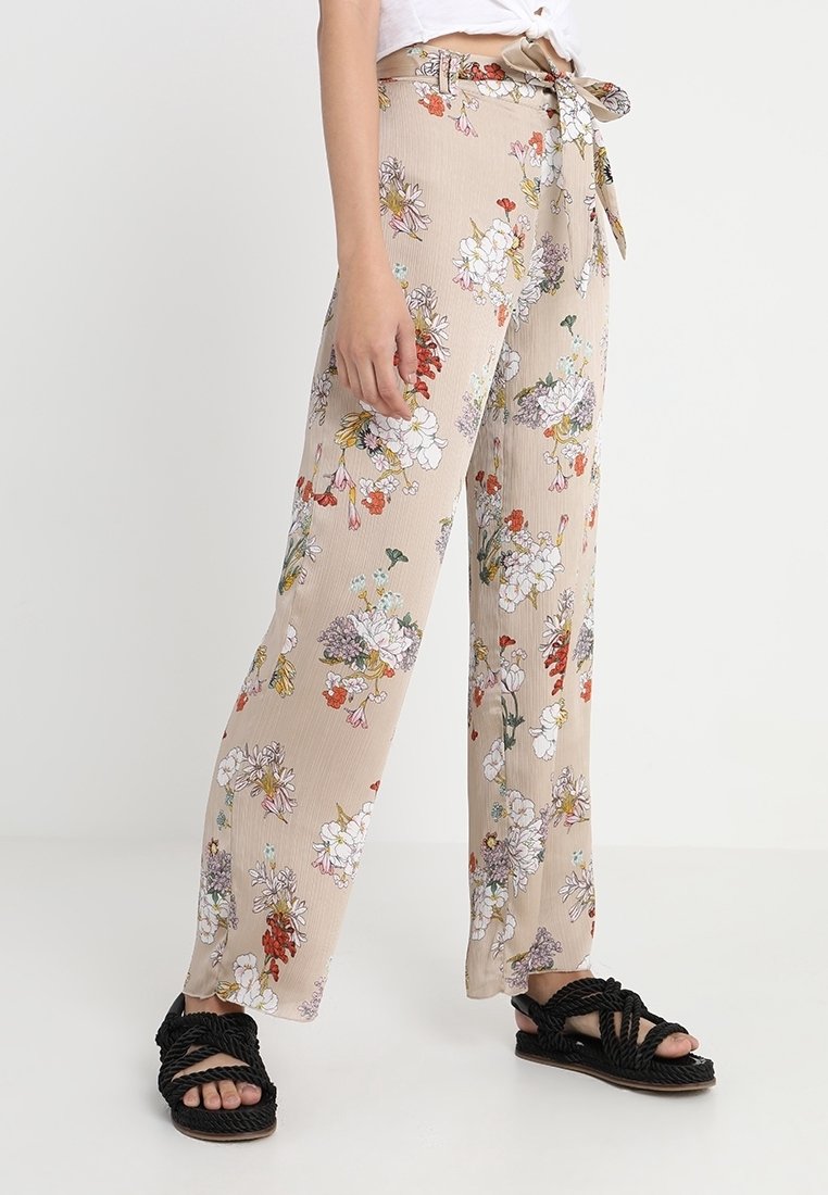 Pantalon large beige à fleurs avec lien sur le côté, porté avec des sandales noires tressées et un crop top blanc noué.