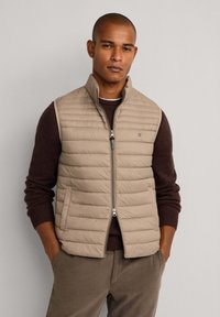 Chaleco de plumas beige con cuello alto, patrón de acolchado horizontal y cremalleras en la parte delantera; llevado sobre un suéter marrón oscuro.