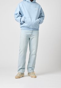 Vaalean sininen huppari, jossa on kenguru-tasku ja logo, yhdistettynä vaaleisiin farkkuihin. Jalkineissa on beigen ja kremin sävyjä.