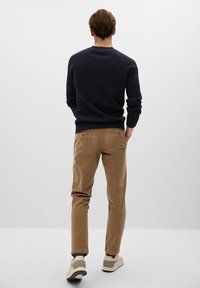 Homme debout, de dos, portant un pull texturé bleu foncé, un pantalon beige, des chaussettes foncées et des baskets beiges sur fond blanc.