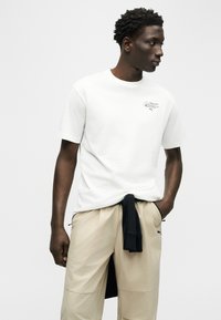 Άνδρας με λευκό t-shirt Puma και μπεζ παντελόνι, με μαύρο ρούχο δεμένο γύρω από τη μέση, κοιτάζει προς τα δεξιά με φόντο απλή επιφάνεια.