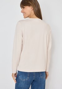 Camicia a maniche lunghe beige chiaro con scollo rotondo, texture morbida e vestibilità casual, abbinata a jeans scuri.