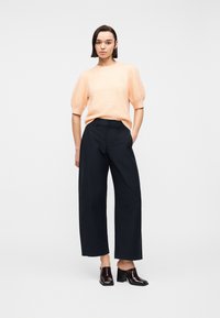 Femme debout, main dans la poche, portant un pull à manches courtes pêche, un pantalon large noir et des mules à talons foncés sur fond blanc.