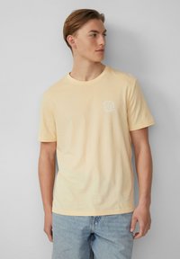 T-shirt en coton jaune clair avec un col rond, des manches courtes et une petite impression graphique sur la poitrine. Texture douce et coupe décontractée.
